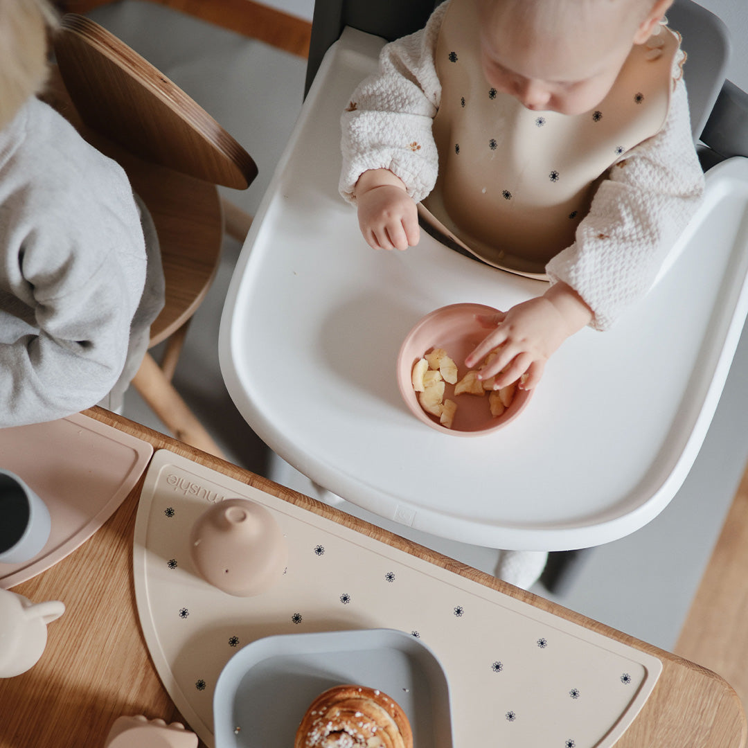 Een baby met een beige siliconen slabbetje eet uit een kommetje in een kinderstoel.