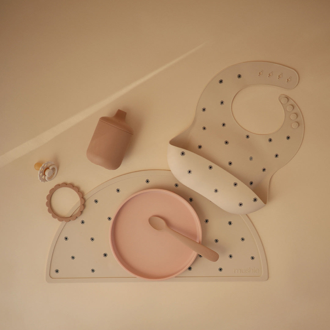Een collectie Mushie babyvoedingsartikelen, waaronder een beige siliconen slabbetje met madeliefjespatroon, een placemat, bord, lepel, fles en speen.