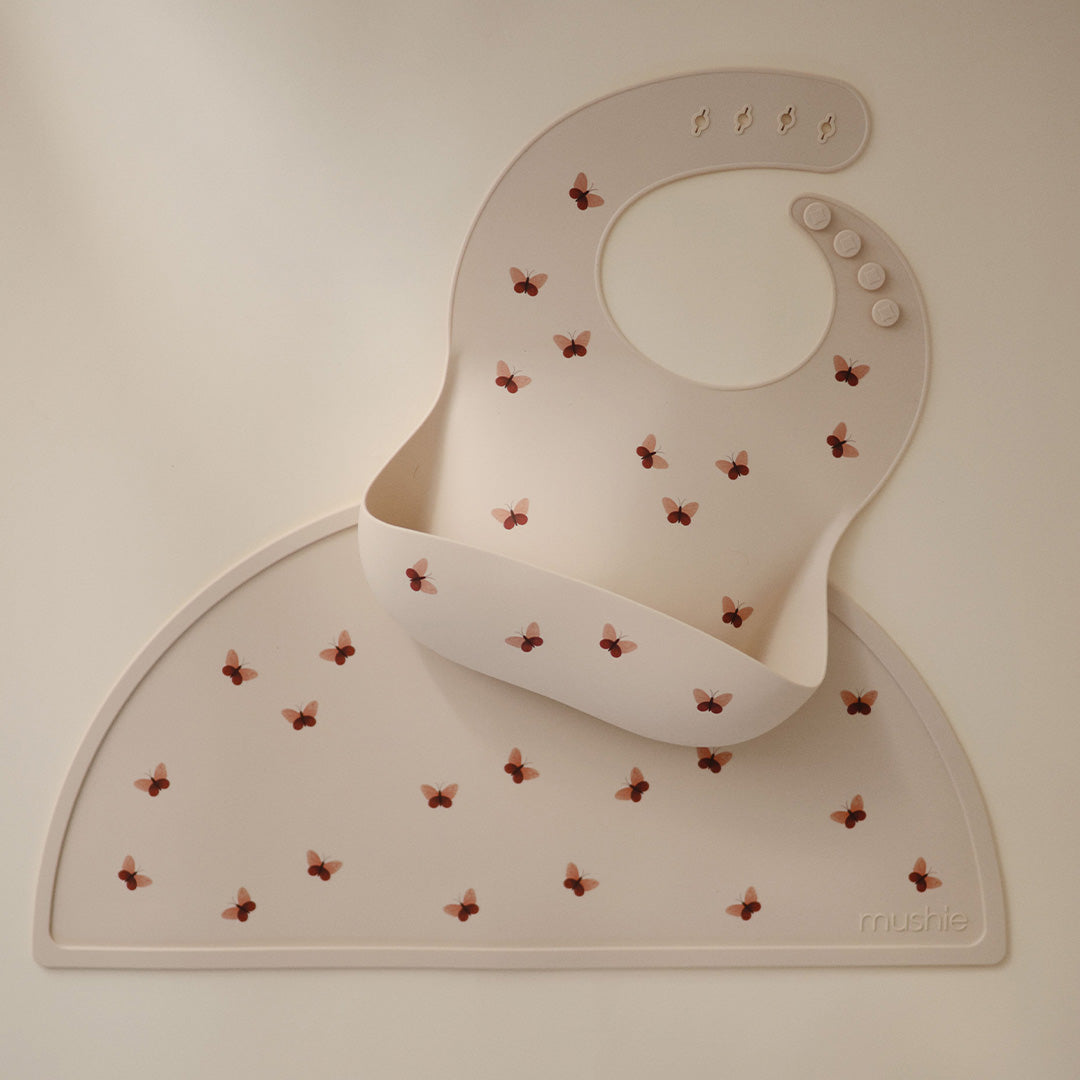 Mushie siliconen slabbetje met vlinders ligt op een bijpassende mushie placemat.