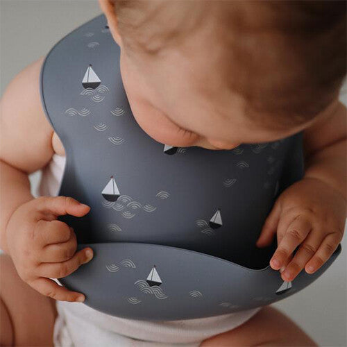 Close-up van een baby die het mushie grijze siliconen slabbetje met zeilbotenpatroon draagt.