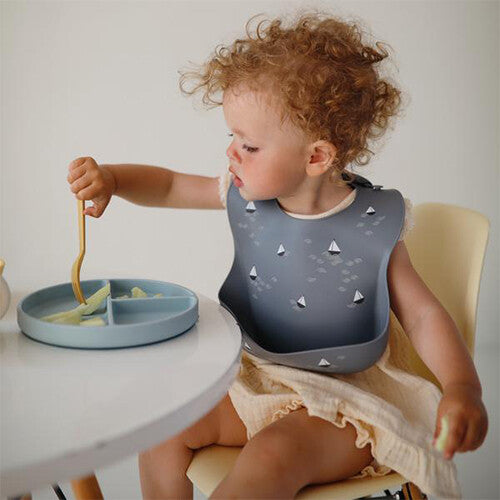 Een peuter met krullen draagt het mushie grijze siliconen slabbetje met zeilbotenpatroon tijdens het eten aan tafel.