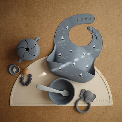 Een collectie mushie babyvoedings- en speelgoedartikelen, waaronder het grijze siliconen slabbetje met zeilbotenpatroon, op een beige mat.