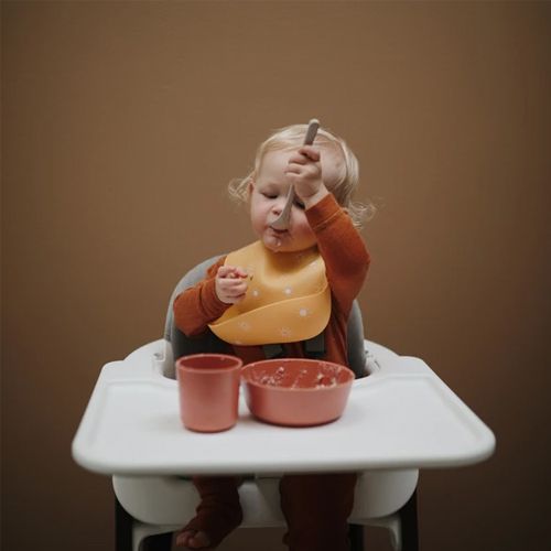 Een baby in een kinderstoel draagt een geel slabbetje en eet met een lepel.