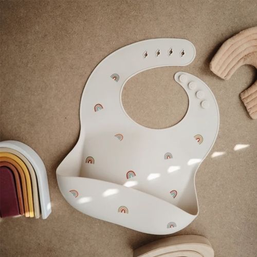 mushie siliconen slabbetje met regenboogpatroon liggend op een bruine ondergrond met houten speelgoed.