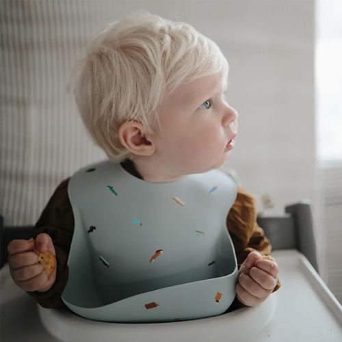 Een baby draagt een lichtblauw mushie siliconen slabbetje met retro auto's in een kinderstoel.