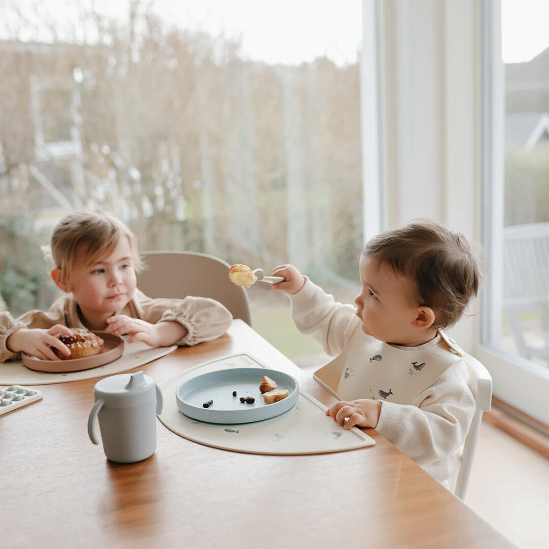 Twee peuters eten aan tafel, waarbij één kind een mushie slabbetje met walvissen draagt.