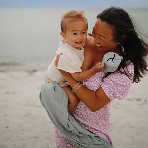 Een lachende vrouw houdt een baby vast op het strand, met de mushie siliconen snackbeker in haar hand.
