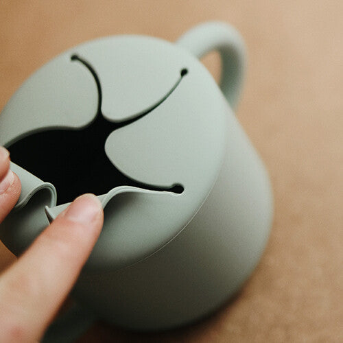 Een vinger duwt door de flexibele siliconen opening van de mushie snackbeker.