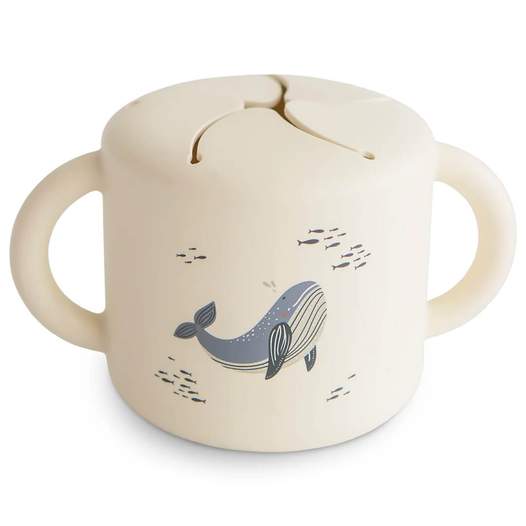 Mushie siliconen snackbeker in beige met walvispatroon, schuin gefotografeerd.