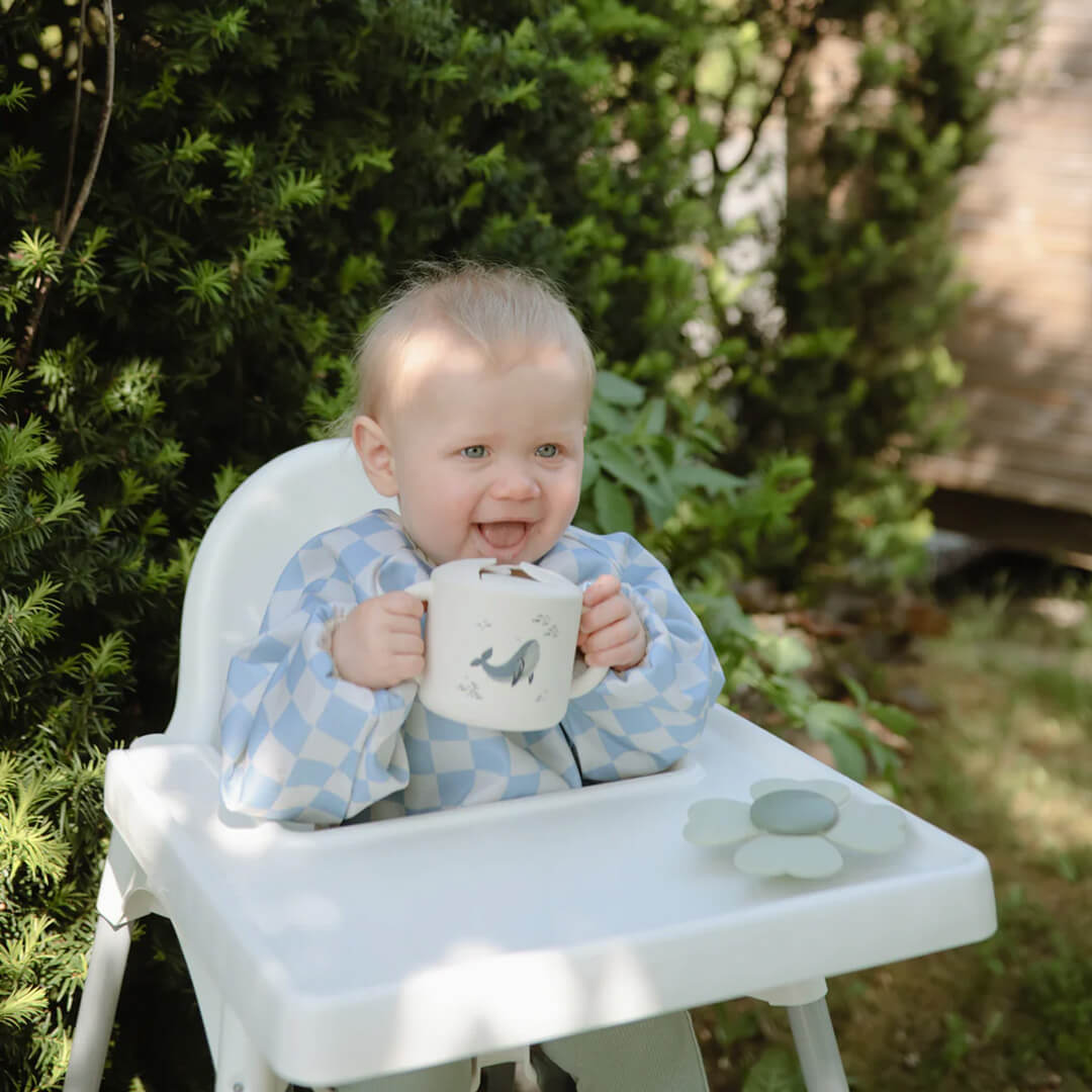 Een lachende baby in een kinderstoel houdt de mushie snackbeker met walvispatroon vast.