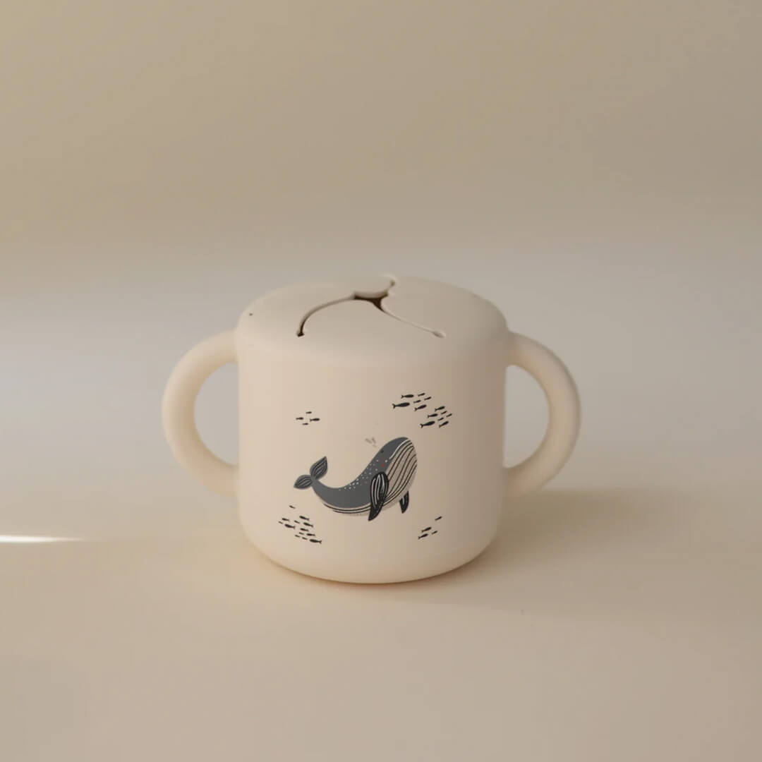 Mushie siliconen snackbeker in beige met walvispatroon, van voren gefotografeerd.