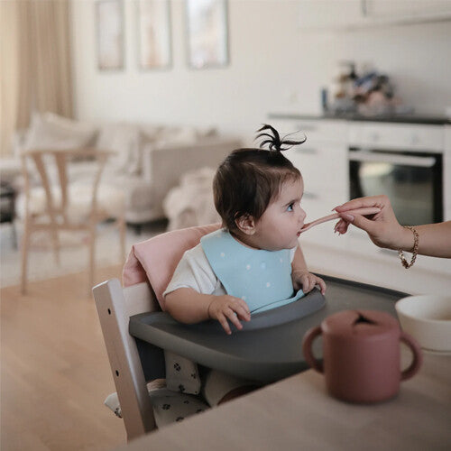 Een baby in een kinderstoel wordt gevoed, met een mushie snackbeker op tafel.