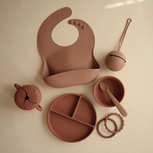 Een collectie mushie babyvoedingsbenodigdheden in woodchuck kleur, waaronder een slab en snackbeker.