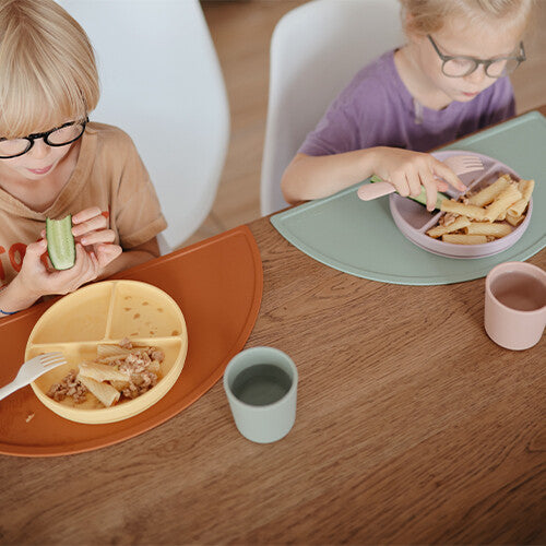 Twee kinderen eten aan tafel met mushie siliconen vakjesborden en placemats.