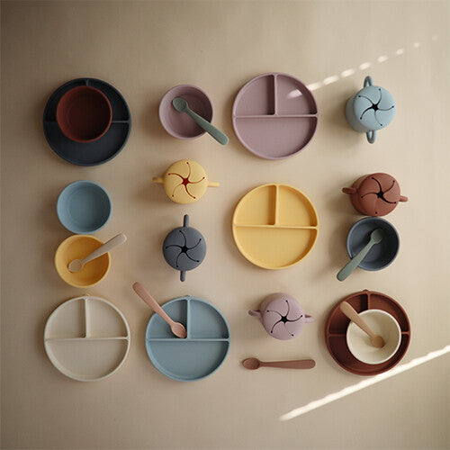 Collectie van mushie siliconen voedingsbenodigdheden zoals vakjesborden, kommen en snackcups in diverse pastelkleuren.