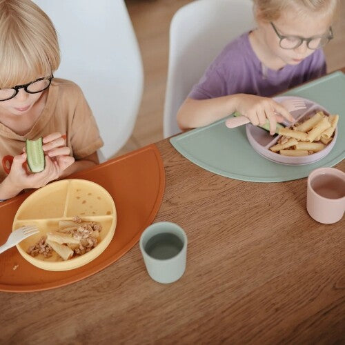 Twee kinderen eten pasta van mushie siliconen vakjesborden aan een houten tafel.