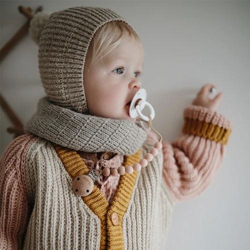Een baby met een Mushie speenketting met roze kralen, bevestigd aan een speen en trui.