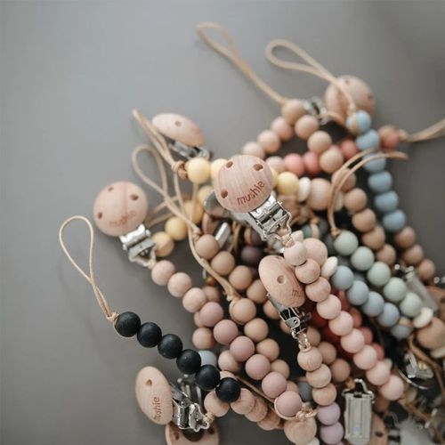 Een collectie Mushie speenkettingen in verschillende kleuren, met houten clips.