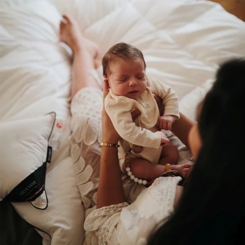 Een slapende pasgeboren baby met een speenketting, vastgehouden door een volwassene op een bed.