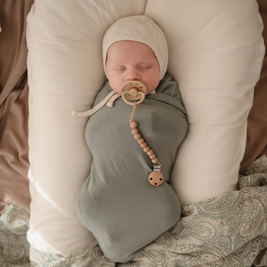 Een slapende baby met een speenkoord en speen, gewikkeld in een groene doek.