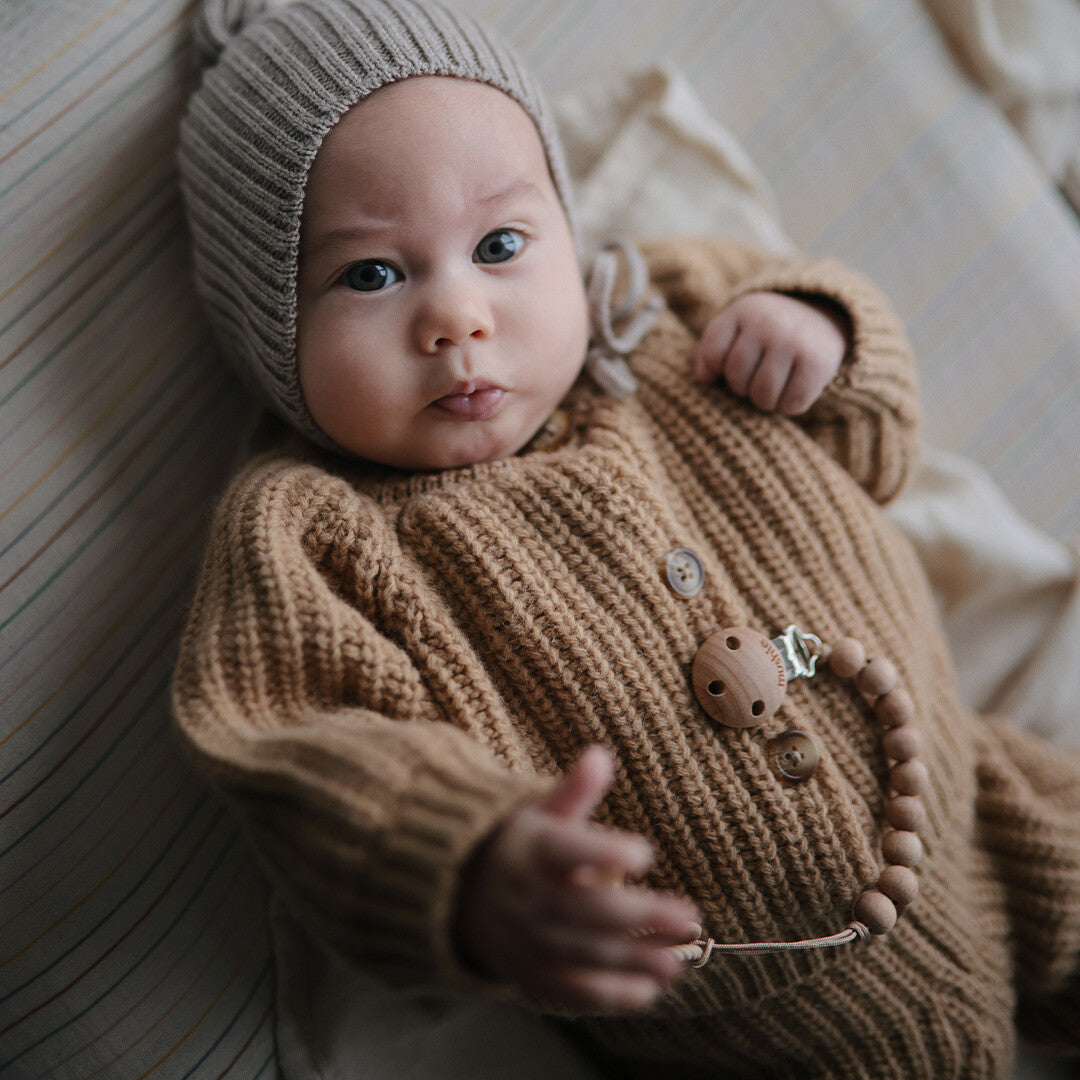 Een baby met een speenkoord, gekleed in een gebreid bruin pakje en een grijze muts.