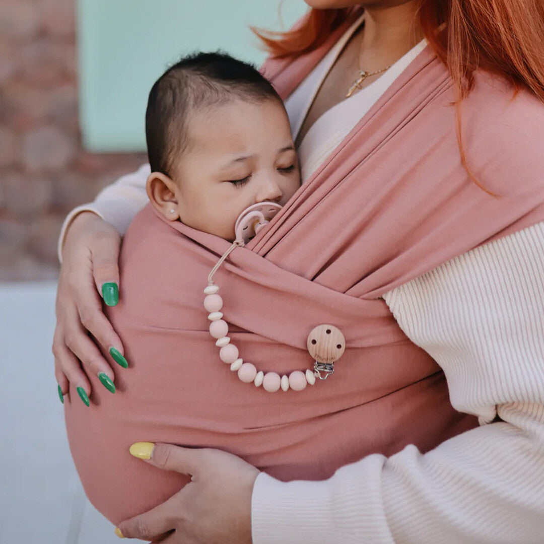 Slapende baby in een roze draagdoek met een Mushie speenkoord en speen.