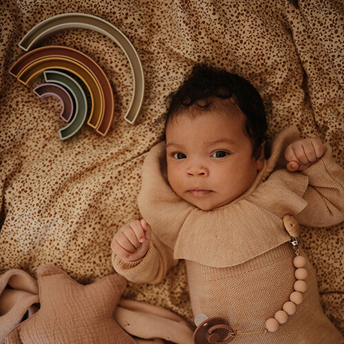 Een baby ligt op een deken met de mushie regenboog stapeltoren ernaast.