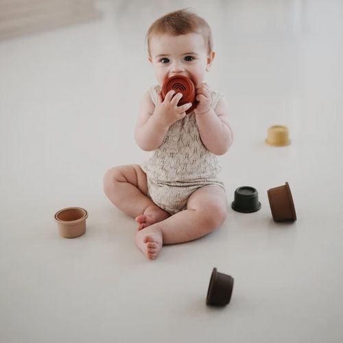 Een baby zit op de grond en bijt op een rode mushie stapelbeker, met andere bekers verspreid eromheen.