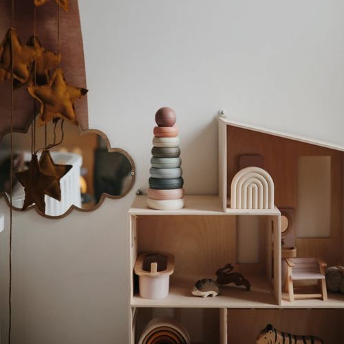 Mushie stapeltoren met veelkleurige ringen op een plank naast een houten poppenhuis.
