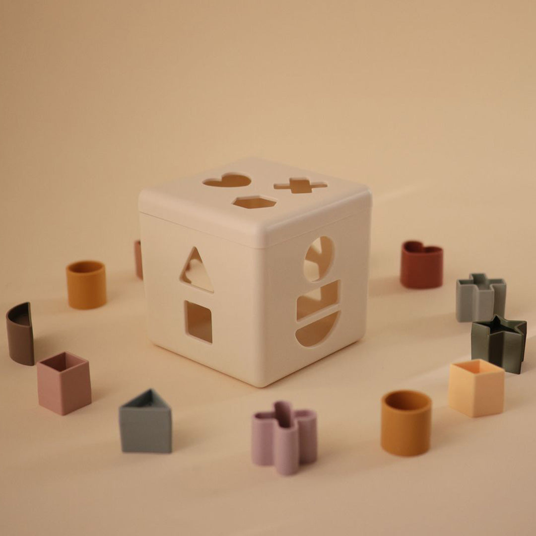 De beige mushie vormendoos staat centraal, omringd door alle gekleurde geometrische vormen op een lichte ondergrond.