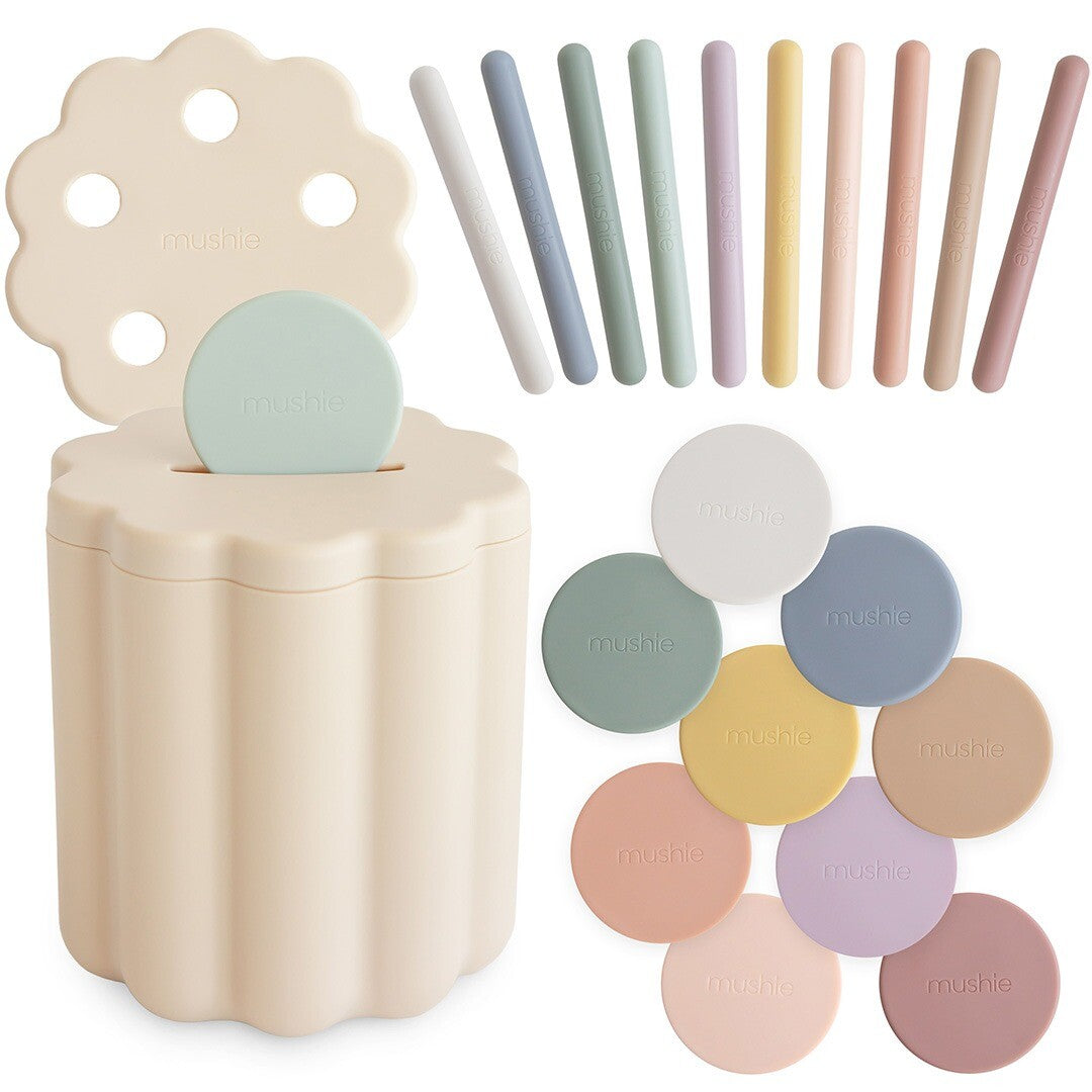 Mushie vormenstoof munt en buis babyspeelgoed set met beige container, deksel, en diverse pastelkleurige munten en staafjes.