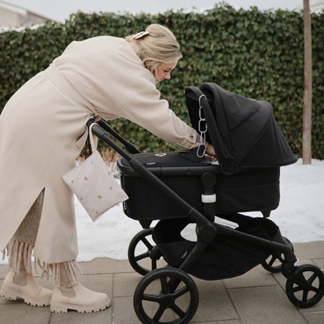 Een vrouw bevestigt een mushie waterdichte opbergzak met hartjespatroon aan een zwarte kinderwagen buiten in de sneeuw.