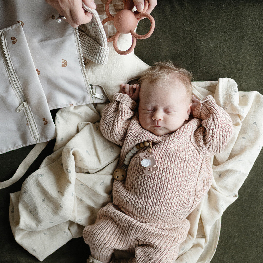 Een slapende baby ligt naast een mushie waterproof opbergzak met regenboogpatroon en een bijtring.