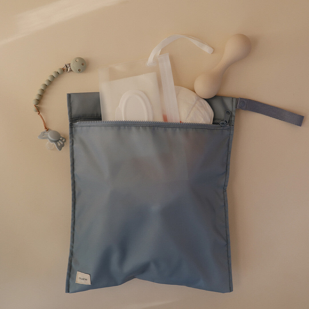 Een open, lichtblauwe mushie waterproof opbergzak gevuld met babyartikelen zoals doekjes en een rammelaar.