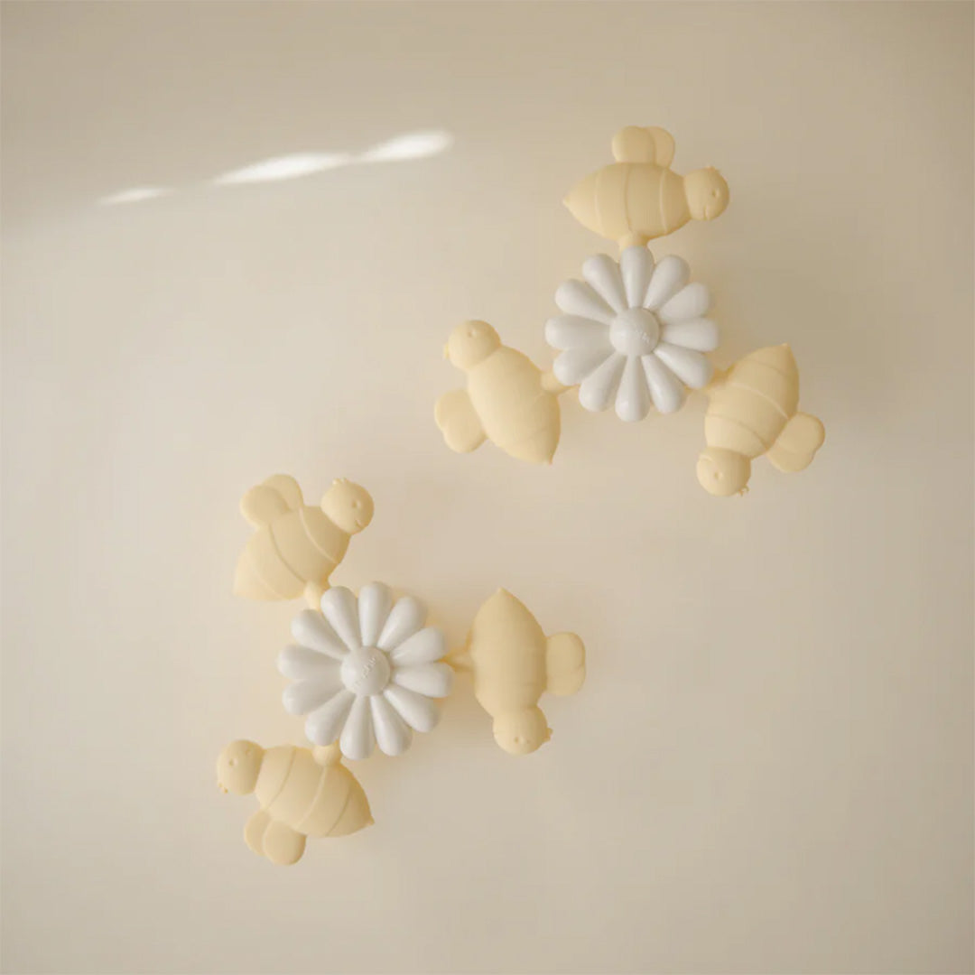 Twee mushie babyspeelgoed spinners met zuignap, elk met een witte bloem en drie beige bijen, liggen op een lichte ondergrond.