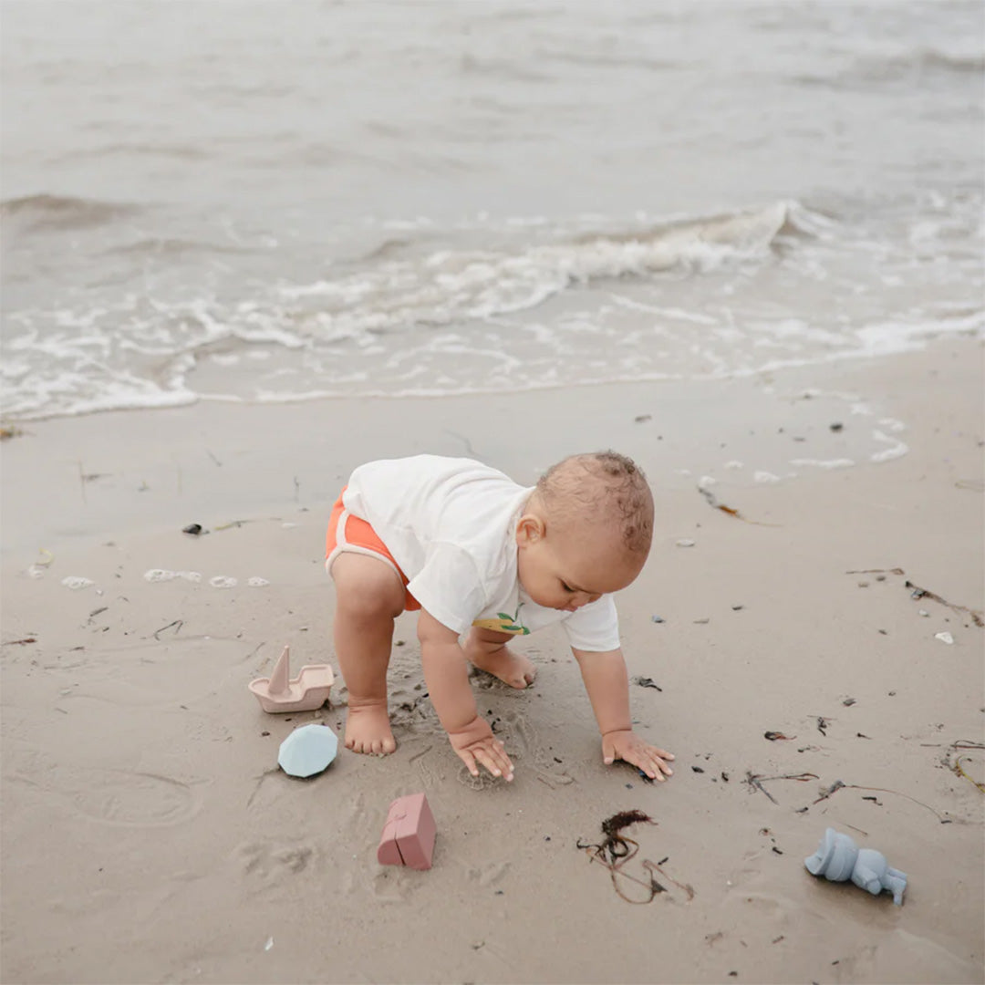Een baby kruipt op het strand met Mushie piraten badspeeltjes om zich heen.