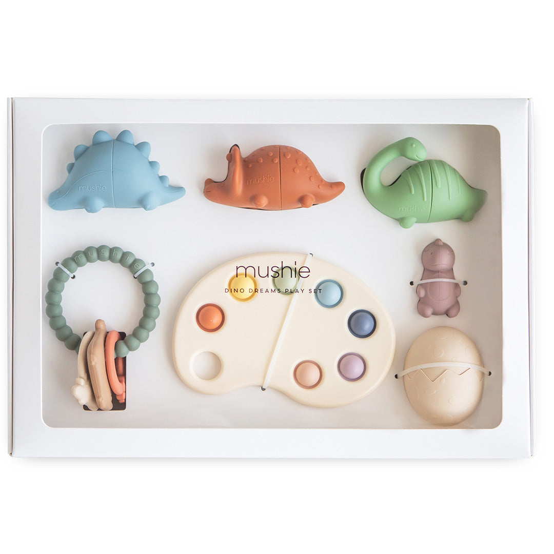 mushie Dino Dreams speelset in een witte verpakking met een kijkvenster, met diverse siliconen babyspeeltjes in dinosaurus-thema.