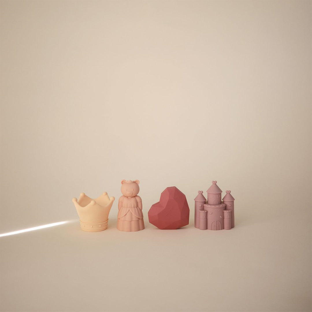 Vier Mushie siliconen babyspeeltjes in roze en beige, waaronder een kroon, beerprinses, hart en kasteel, op een lichte ondergrond.