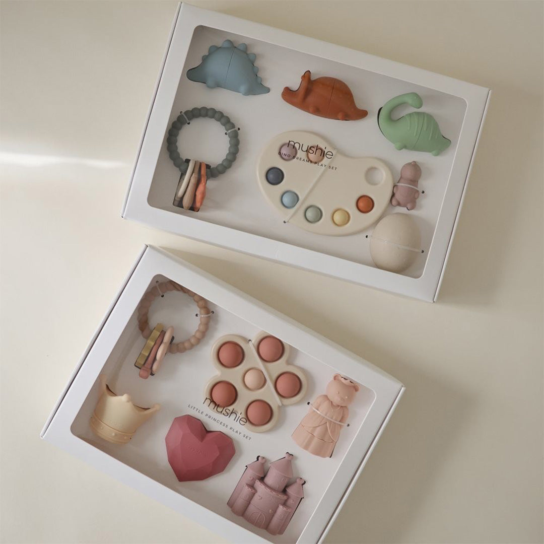 Twee Mushie geschenkdozen met siliconen babyspeelgoed, waaronder de Little Princess en Dino Play Set, schuin van bovenaf gefotografeerd.
