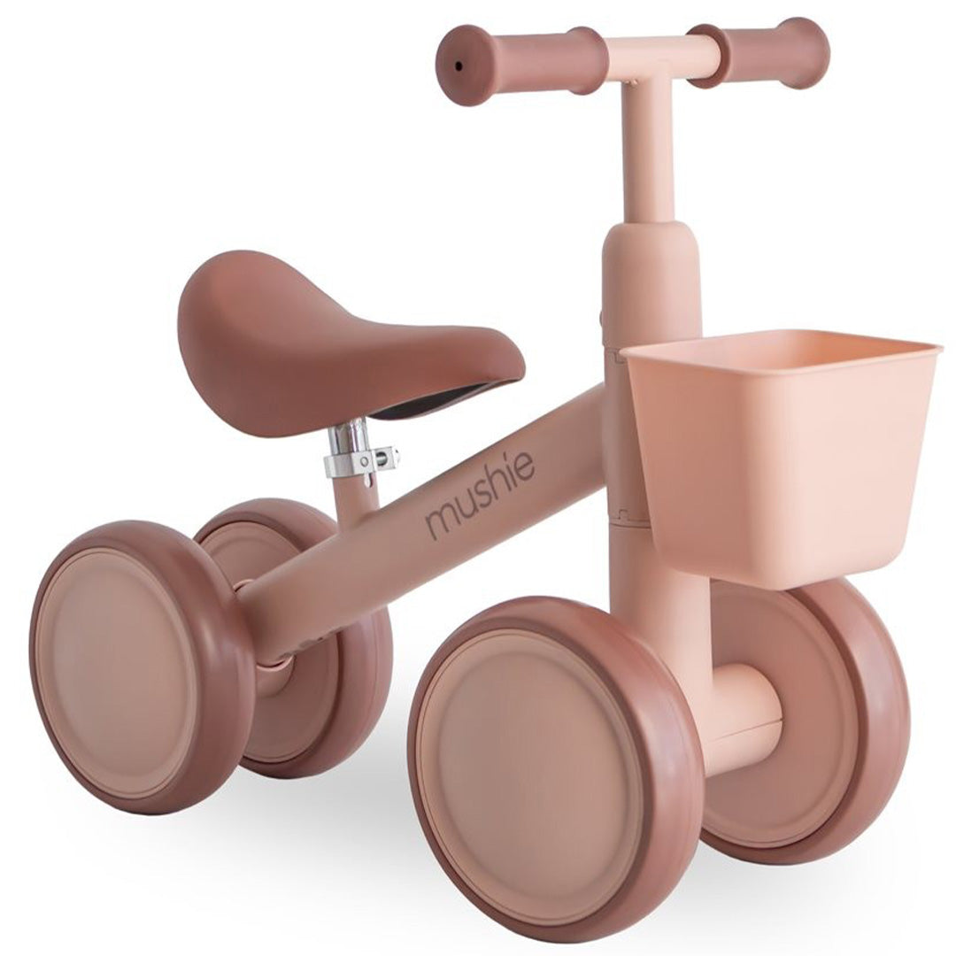 Mushie loopfiets voor peuters in blush roze met vier wielen en een mandje, op een witte achtergrond.