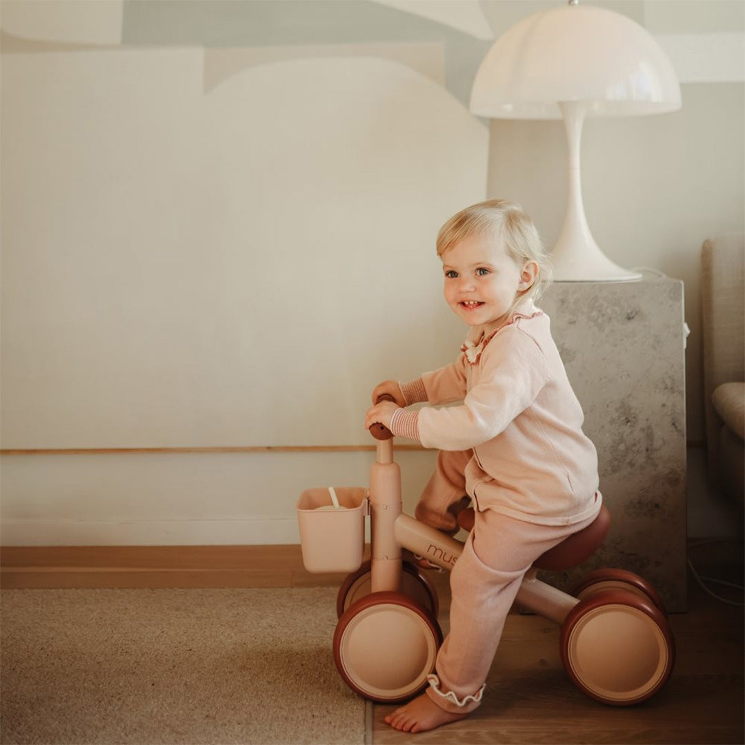 Een lachende peuter zit op een Mushie loopfiets in blush roze, binnenshuis.