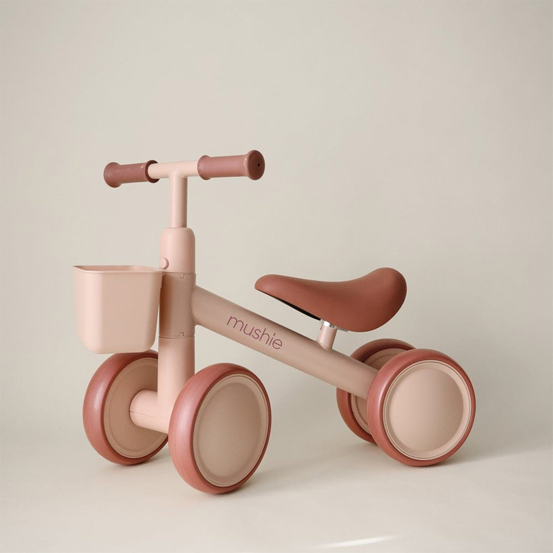 Mushie loopfiets voor peuters in blush roze met vier wielen en een mandje, op een beige achtergrond.