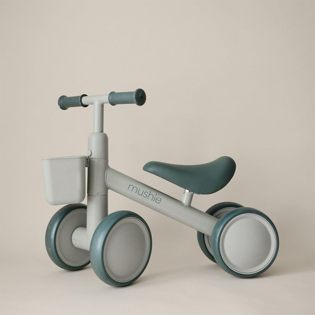Mushie loopfiets voor peuters in saliegroen met een mandje aan het stuur, tegen een lichte achtergrond.