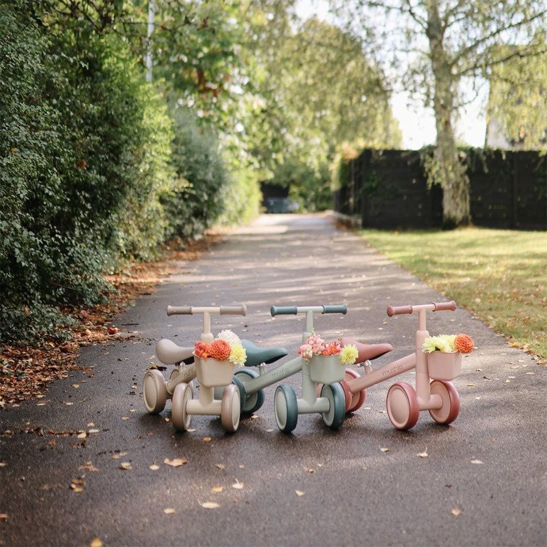 Drie Mushie loopfietsen in beige, saliegroen en roze met bloemen in de mandjes, staand op een pad buiten.