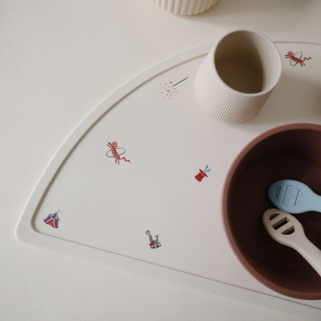 Detail van de beige baby placemat met circusfiguren, met een beker en kom erop.
