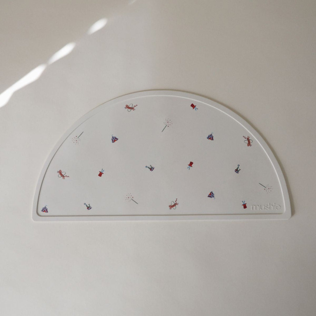 mushie baby placemat in beige met circusfiguren, liggend op een lichte ondergrond.