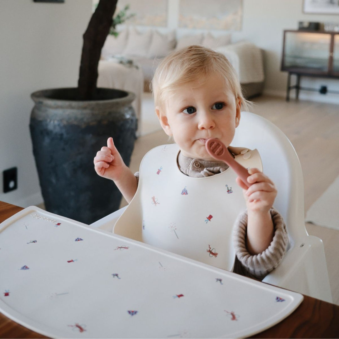 Een baby in een kinderstoel draagt een beige siliconen slabbetje met circusfiguren en eet met een lepel.