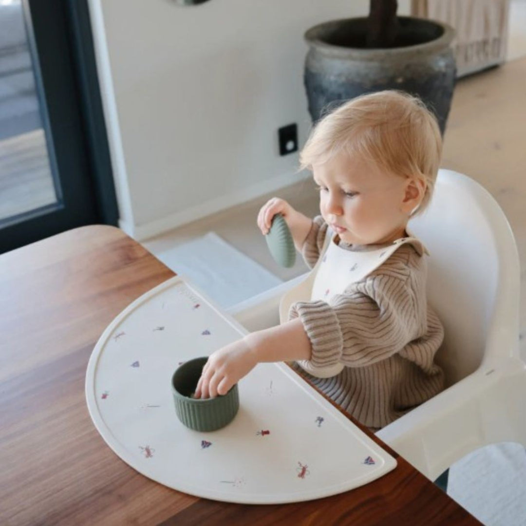 Een baby in een kinderstoel draagt een beige siliconen slabbetje met circusfiguren en speelt met een kommetje.