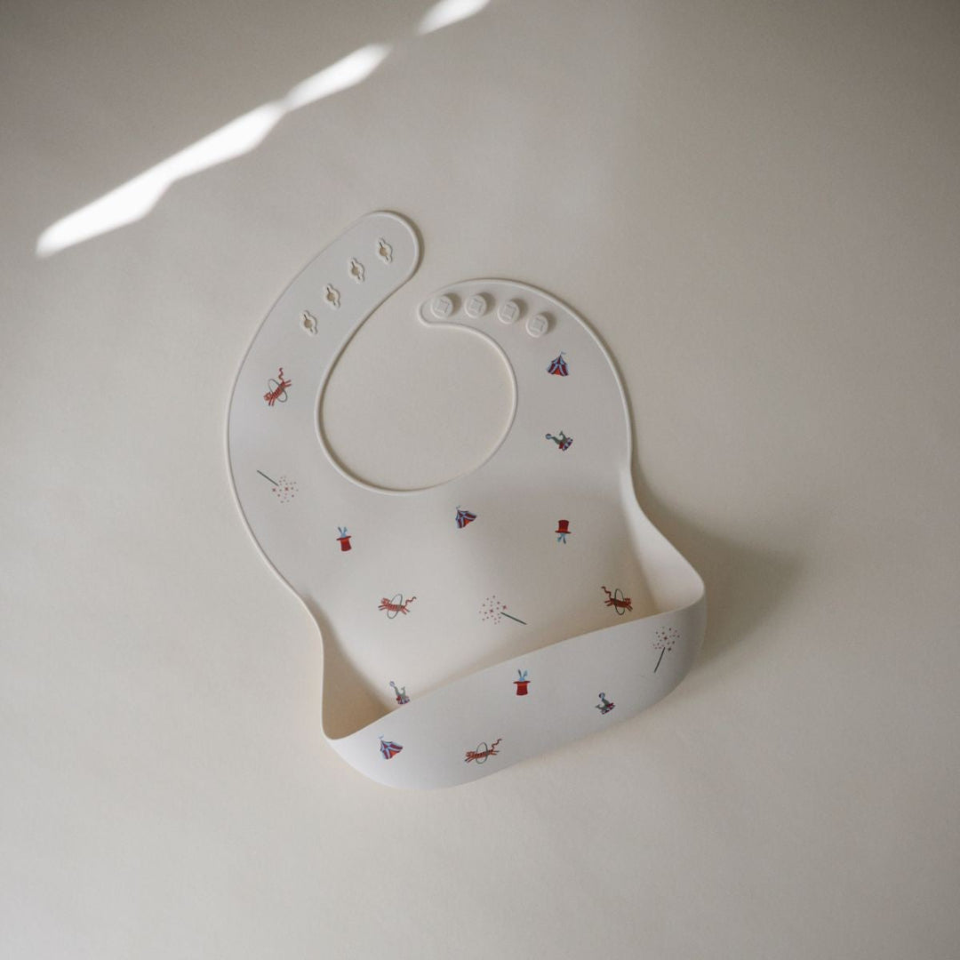 Mushie siliconen babyslabbetje in beige met circusfiguren, van bovenaf gezien.