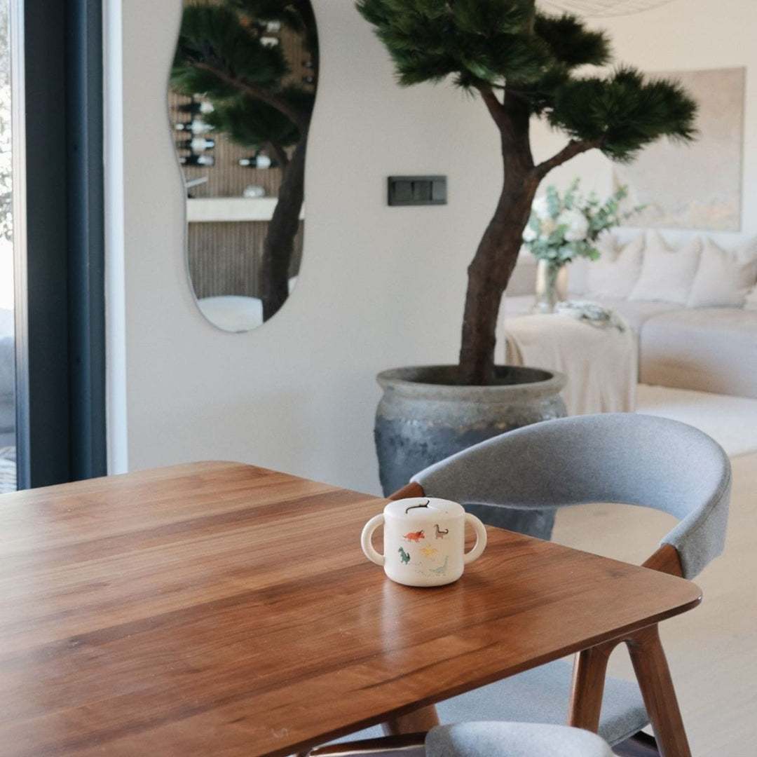 Een mushie siliconen snackbeker met dinosauruspatronen staat op een houten tafel in een modern interieur.